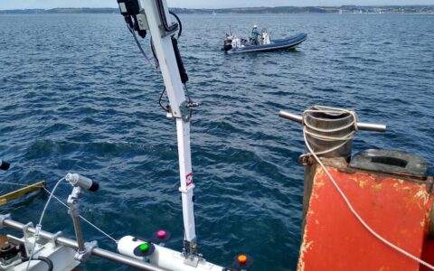 Photograph of Proteus 100 robotic arm lifting model AUV out of the sea with support boat in background.